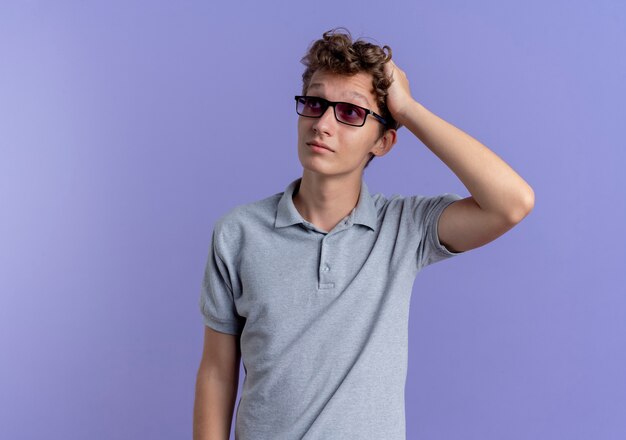 Joven de gafas negras con camisa de polo gris mirando a un lado con la mano en la cabeza confundida de pie sobre la pared azul