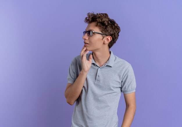 Joven de gafas negras con camisa de polo gris mirando a un lado perplejo parado sobre la pared azul