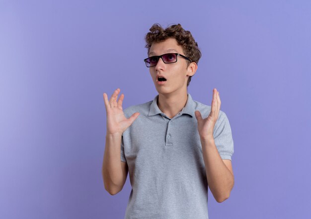 Joven de gafas negras con camisa de polo gris mirando a un lado sorprendido y asombrado con las manos levantadas sobre la pared azul