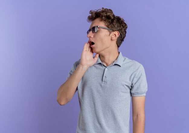 Joven de gafas negras con camisa de polo gris mirando a un lado sorprendido y asombrado de pie sobre la pared azul