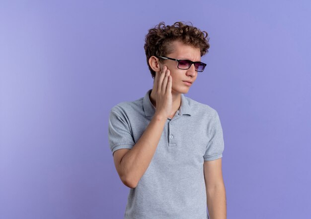 Joven de gafas negras con camisa de polo gris mirando a un lado tocando su mejilla sintiendo dolor de muelas de pie sobre la pared azul