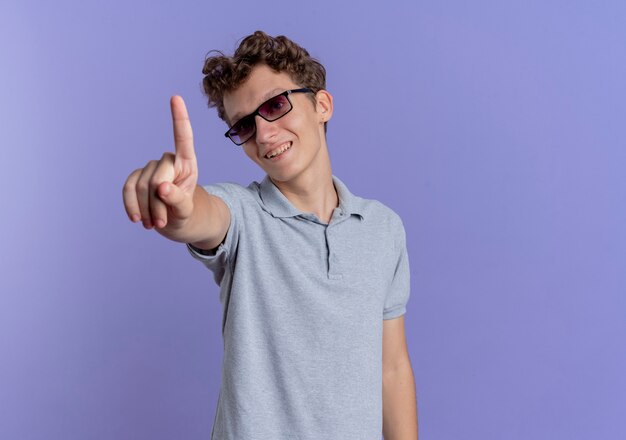 Joven de gafas negras con camisa polo gris mostrando el dedo índice sonriendo con cara feliz de pie sobre la pared azul