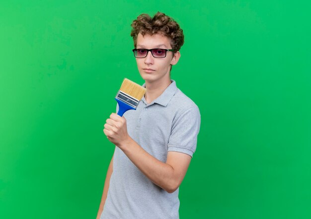 Joven de gafas negras con camisa de polo gris con pincel mirando a la cámara con cara de escéptico serio sobre verde