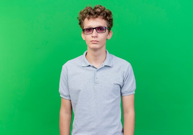 Joven de gafas negras con camisa de polo gris con rostro serio de pie sobre la pared verde