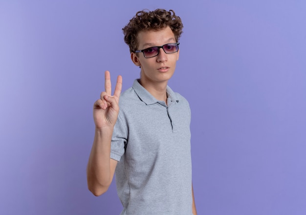 Joven de gafas negras con camisa de polo gris con rostro seriosu mostrando y apuntando hacia arriba con los dedos número dos sobre azul