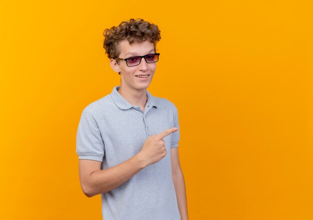 Joven de gafas negras con camisa de polo gris sonriendo alegremente apuntando con el dedo índice hacia el lado de pie sobre la pared naranja