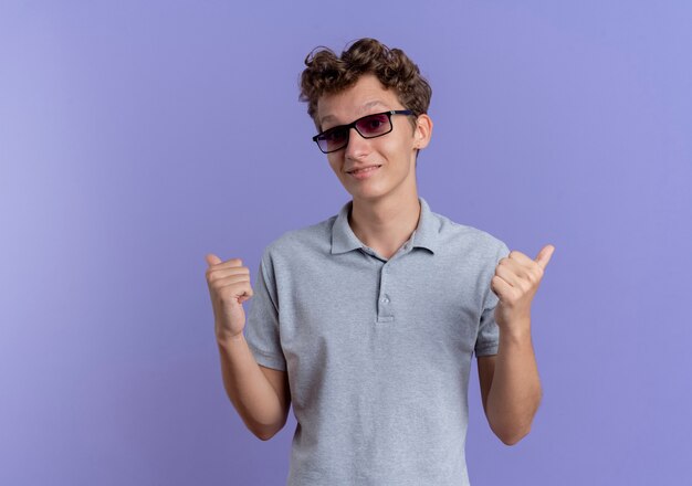 Joven de gafas negras con camisa de polo gris sonriendo apuntando hacia atrás con los pulgares de pie sobre la pared azul