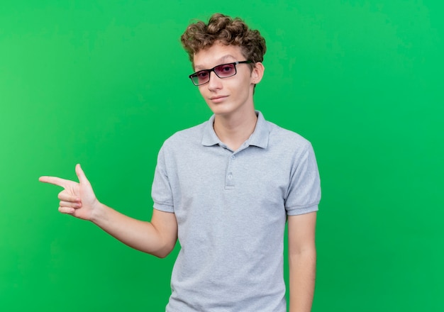 Joven de gafas negras con camisa de polo gris sonriendo apuntando con el dedo índice hacia el lado de pie sobre la pared verde