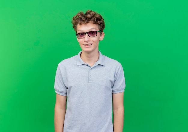 Joven de gafas negras con camisa de polo gris con una sonrisa escéptica de pie sobre la pared verde