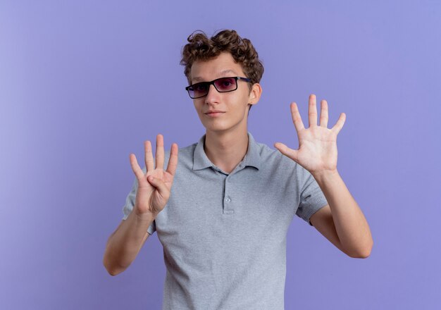 Joven de gafas negras con camisa de polo gris con sonrisa mostrando y apuntando hacia arriba con los dedos número nueve de pie sobre la pared azul