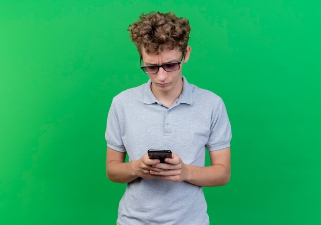 Joven de gafas negras con camisa de polo gris sosteniendo smartphone mirándolo con cara seria sobre verde