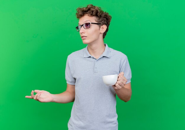 Joven de gafas negras con camisa de polo gris sosteniendo la taza de café mirando a un lado confundido de pie sobre la pared verde