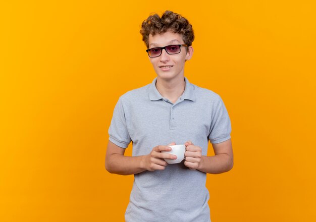 Joven de gafas negras con camisa de polo gris sosteniendo la taza de café sonriendo alegremente sobre verde