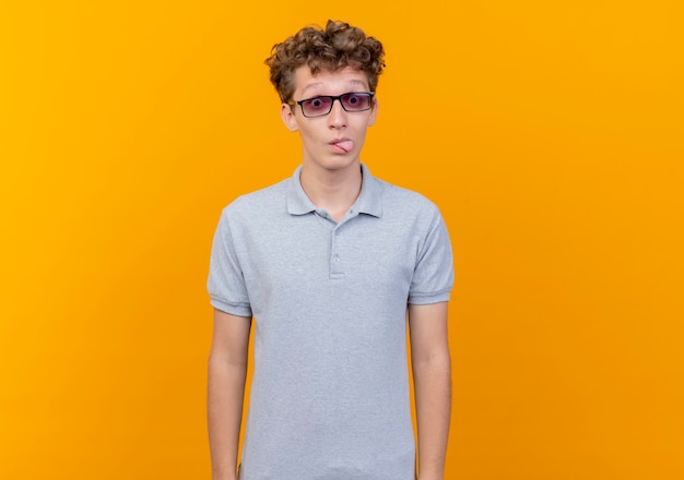 Joven de gafas negras vistiendo polo gris feliz y positivo sacando la lengua sobre naranja