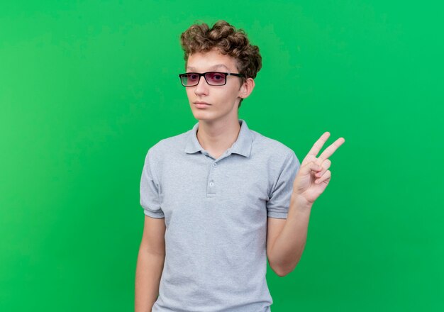 Joven de gafas negras vistiendo polo gris sonriendo mostrando signo v sobre verde