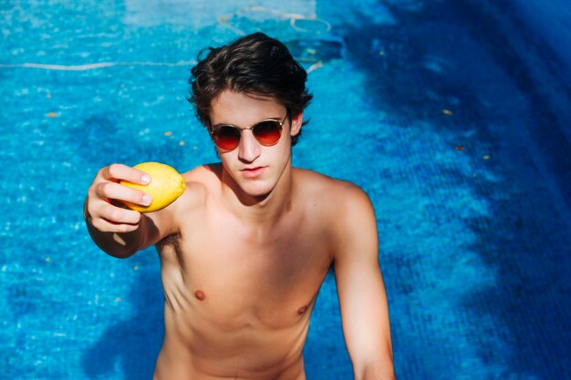 Joven con gafas de sol mostrando limón en la piscina