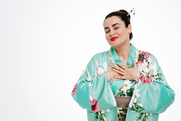 Joven geisha en kimono japonés tradicional feliz y complacida con los ojos cerrados sintiéndose agradecida tomados de la mano en el pecho de pie sobre una pared blanca