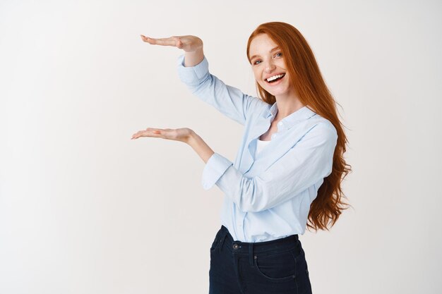 Joven gerente femenina pelirroja mostrando un objeto de gran tamaño, demostrando un producto de gran tamaño y sonriendo, de pie sobre una pared blanca