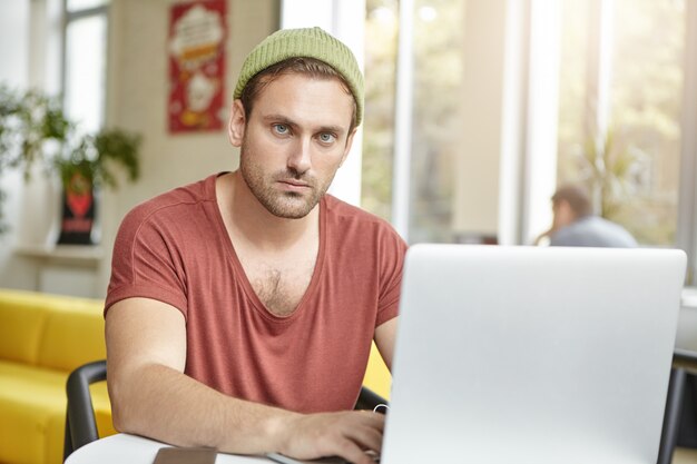 El joven gerente próspero viste una camiseta informal y un sombrero, trabaja en una computadora portátil genérica