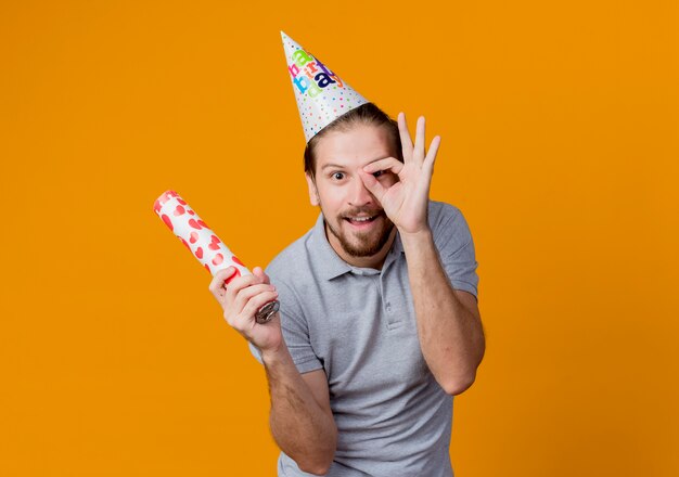 Joven en gorra de vacaciones mostrando signo ok sonriendo alegremente concepto de fiesta de cumpleaños de pie sobre la pared naranja