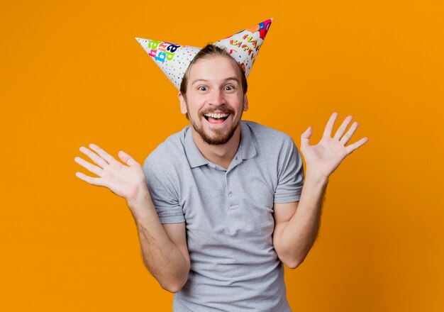 Joven con gorras de vacaciones sonriendo feliz y emocionado concepto de fiesta de cumpleaños de pie sobre la pared naranja