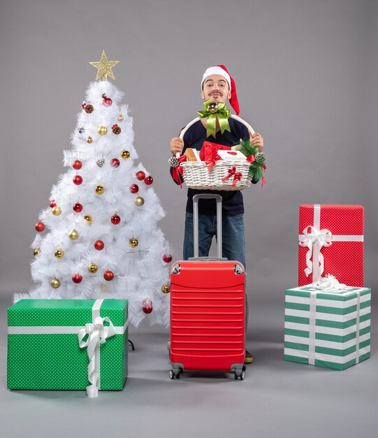 Joven con gorro de Papá Noel sosteniendo una canasta de regalo con ambas manos cerca del árbol de Navidad en gris