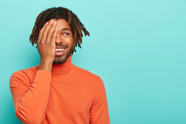 Joven gracioso de piel oscura, cubre la mitad de la cara con la palma de la mano, se ríe positivamente, tiene una sonrisa agradable, usa un jersey de cuello alto naranja