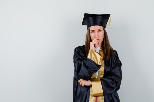 Foto gratuita joven graduada de pie en pose de pensamiento en traje académico y mirando pensativo. vista frontal.