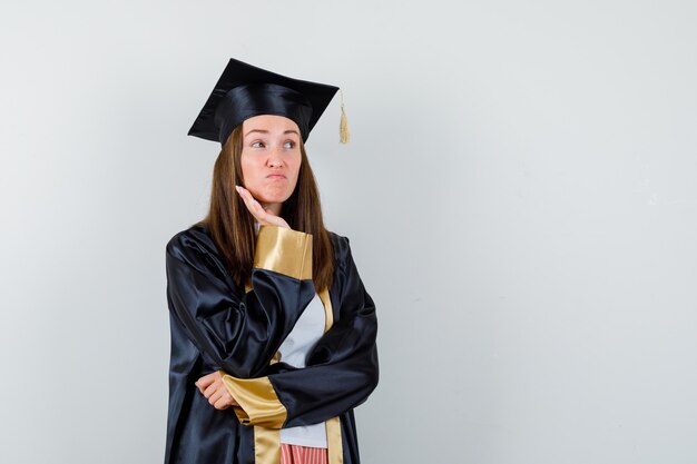 Joven graduada sosteniendo la mano en la mejilla en traje académico y mirando vacilante, vista frontal.