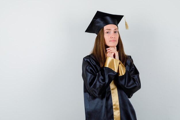 Joven graduada en traje académico cogidos de la mano debajo de la barbilla y mirando esperanzado, vista frontal.