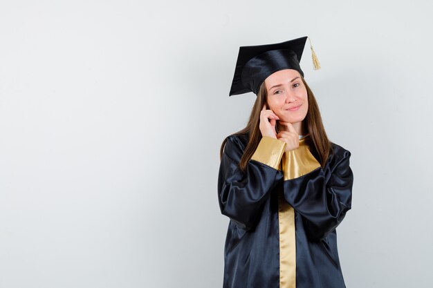 Joven graduada en traje académico de pie mientras posa y mira encantadora, vista frontal.