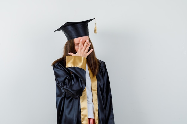 Foto gratuita joven graduada en traje académico sosteniendo la mano en la cara y mirando molesto, vista frontal.