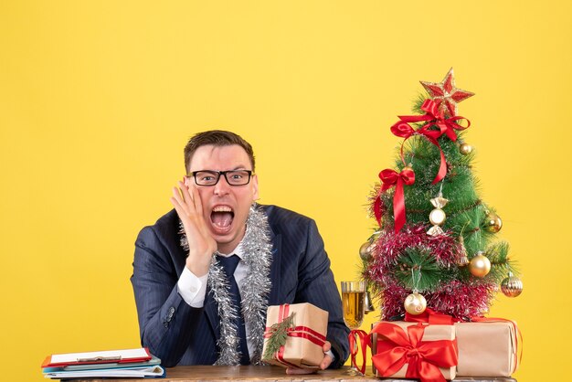 Joven gritando a alguien sentado en la mesa cerca del árbol de Navidad y presenta en amarillo