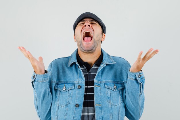 Joven gritando mientras levanta las manos en camiseta, chaqueta, gorra y luce feliz, vista frontal.