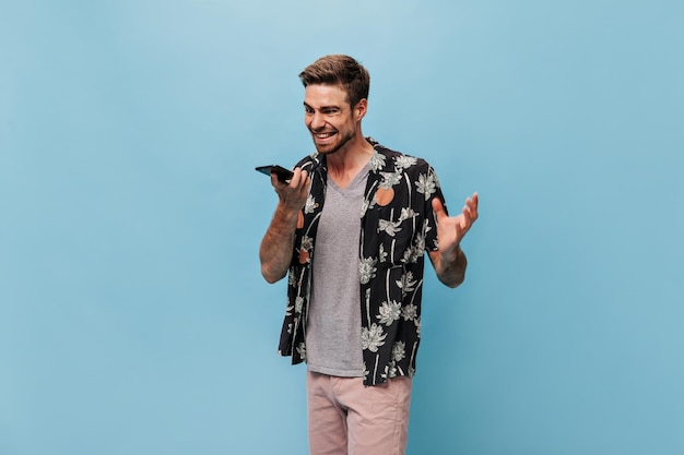 Joven gruñón con camisa negra de moda, camiseta gris y pantalones cortos modernos, habla enojado por teléfono con un fondo azul aislado