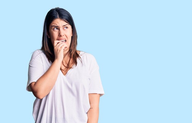 Joven guapa morena con camiseta casual que parece estresada y nerviosa con las manos en la boca mordiéndose las uñas problema de ansiedad