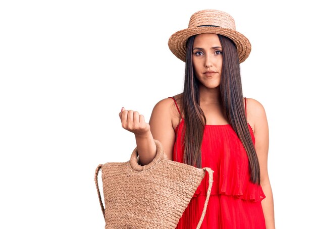 Foto gratuita joven guapa morena con sombrero de verano y vestido sosteniendo una bolsa con actitud de pensamiento y expresión sobria que parece segura de sí misma