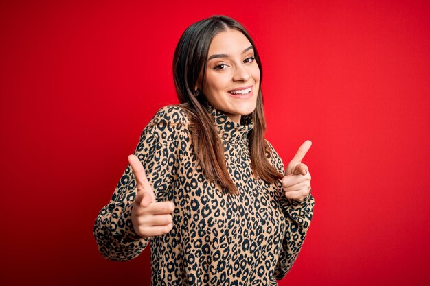 Joven guapa morena con suéter casual de pie sobre fondo rojo señalando con el dedo a la cámara con cara feliz y divertida Buena energía y vibraciones