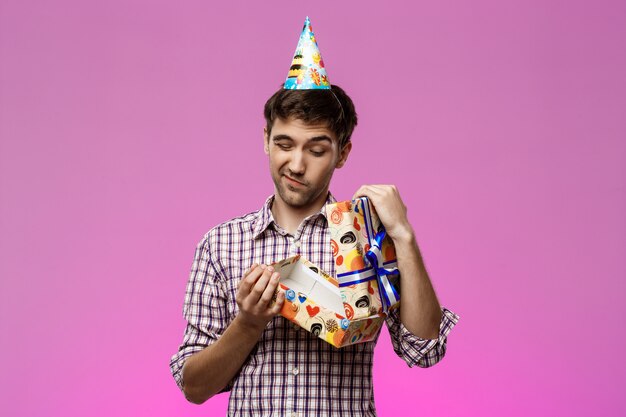 Joven guapo abriendo regalo de cumpleaños sobre pared púrpura.