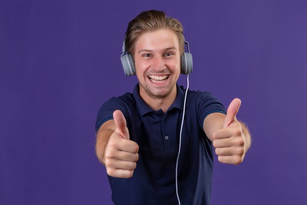 Joven guapo con auriculares mirando a la cámara salió y feliz mostrando los pulgares para arriba sonriendo alegremente sobre fondo púrpura