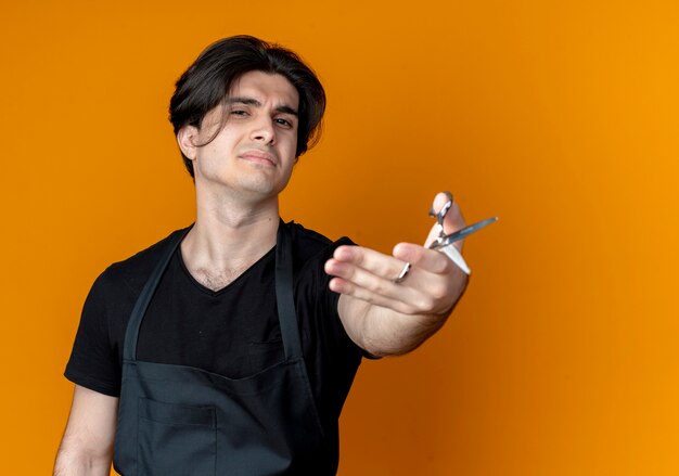 Joven guapo barbero en uniforme sosteniendo unas tijeras aislado en la pared naranja