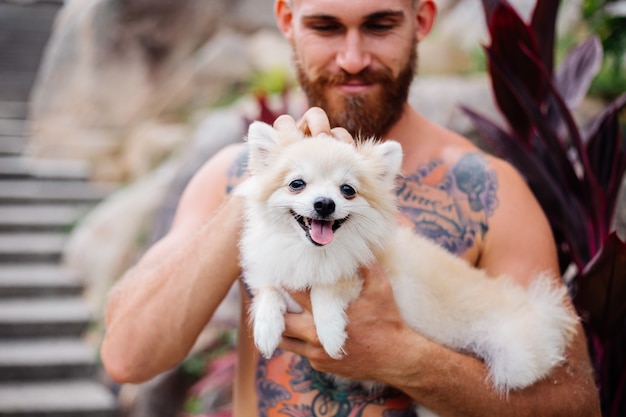 Joven guapo barbudo brutal tatuado feliz sostiene pomerania spitz jugando con mascota encantadora