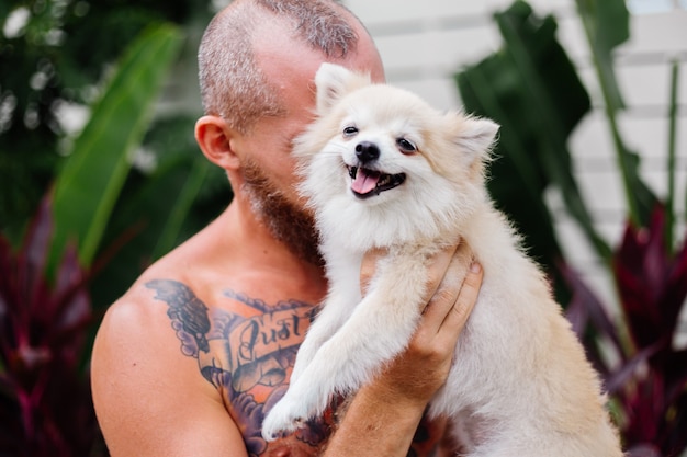 Joven guapo barbudo brutal tatuado feliz sostiene pomerania spitz jugando con mascota encantadora