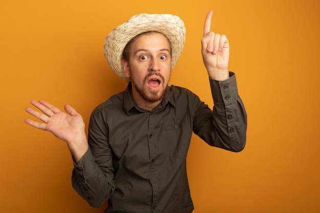 Joven guapo con camisa gris y sombrero de verano asombrado y sorprendido mostrando índice figner y presentando algo con brazo