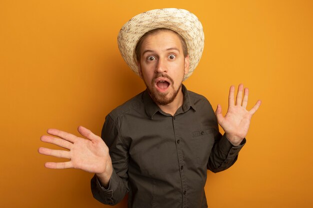 Joven guapo con camisa gris y sombrero de verano sorprendido y asombrado con los brazos levantados