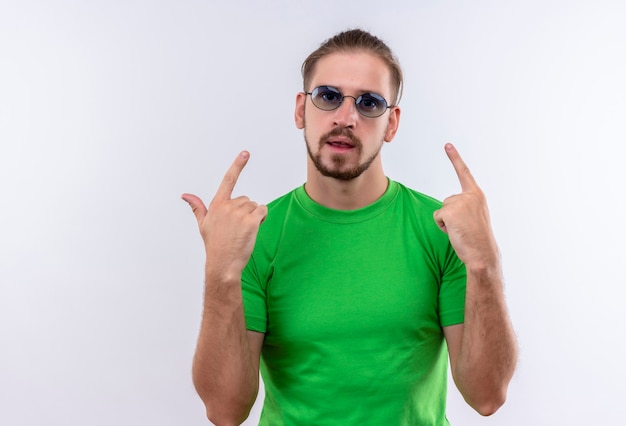 Joven guapo en camiseta verde con gafas apuntando con los dedos hacia arriba mirando confiado de pie sobre fondo blanco.