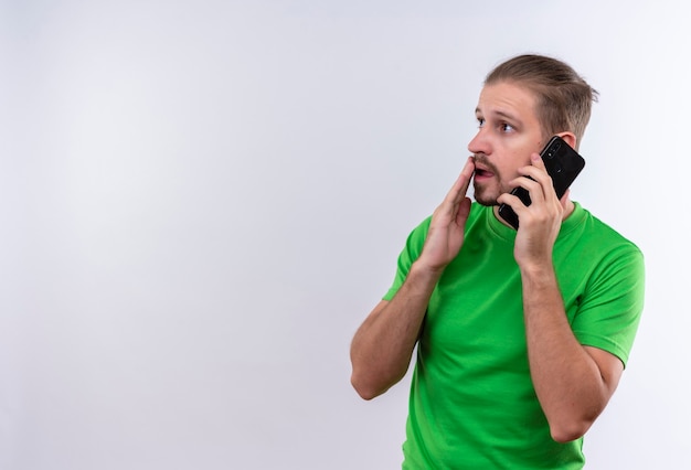 Foto gratuita joven guapo en camiseta verde hablando por teléfono móvil mirando sorprendido y asombrado de pie sobre fondo blanco.