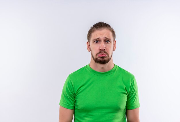 Joven guapo en camiseta verde mirando a la cámara con expresión triste en la cara de pie sobre fondo blanco.