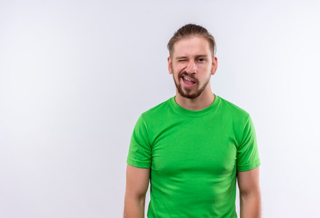 Joven guapo en camiseta verde mirando a cámara haciendo muecas de pie sobre fondo blanco.