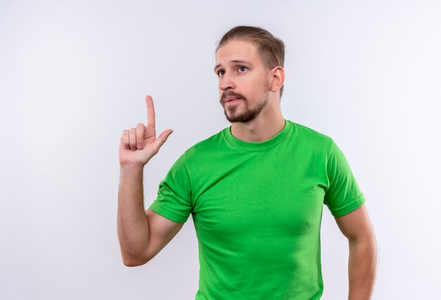 Joven guapo en camiseta verde mirando a un lado apuntando con el dedo hacia arriba mirando confiado tener una idea de pie sobre fondo blanco.